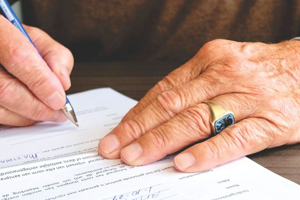 La mano de una persona mayor firmando un documento, con anillo de bodas